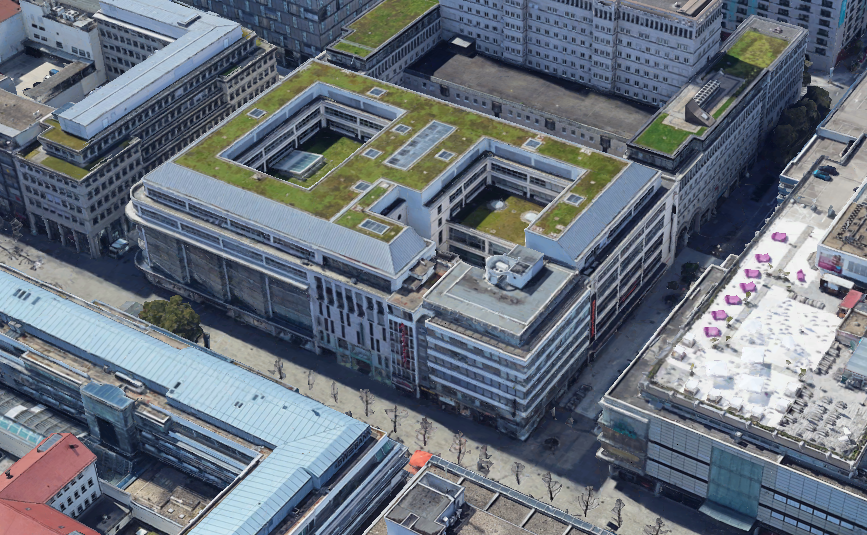 Schrägansicht eines großen, U‑förmigen Gebäudeblocks mit begrünten Dachflächen in dicht bebauter Innenstadtlage.