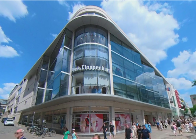 Moderne Straßenperspektive eines mehrgeschossigen Gebäudes mit großer Glasfassade und Passanten im Vordergrund.